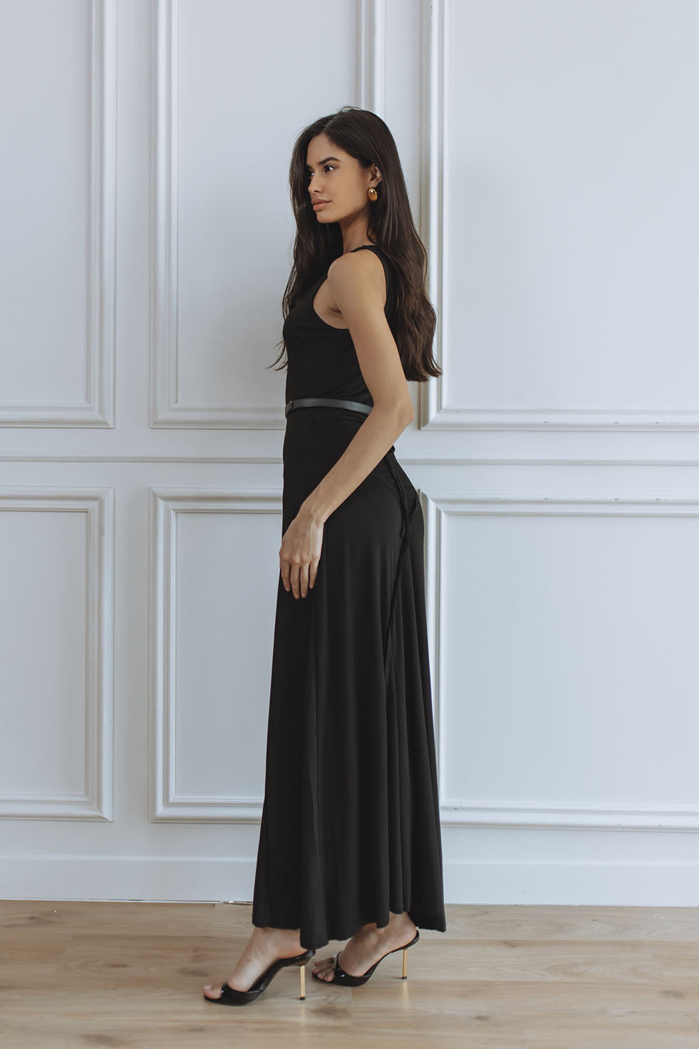A woman modeling a sleeveless black maxi dress with a thin waistband, standing in a room with white paneled walls and light wood flooring.