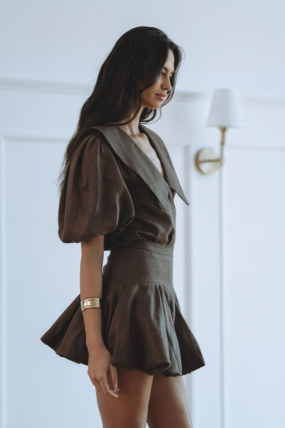 A woman wearing a dark brown short puff-sleeve dress with a wide collar and a flared skirt, standing in a bright white room with a wall lamp visible in the background.