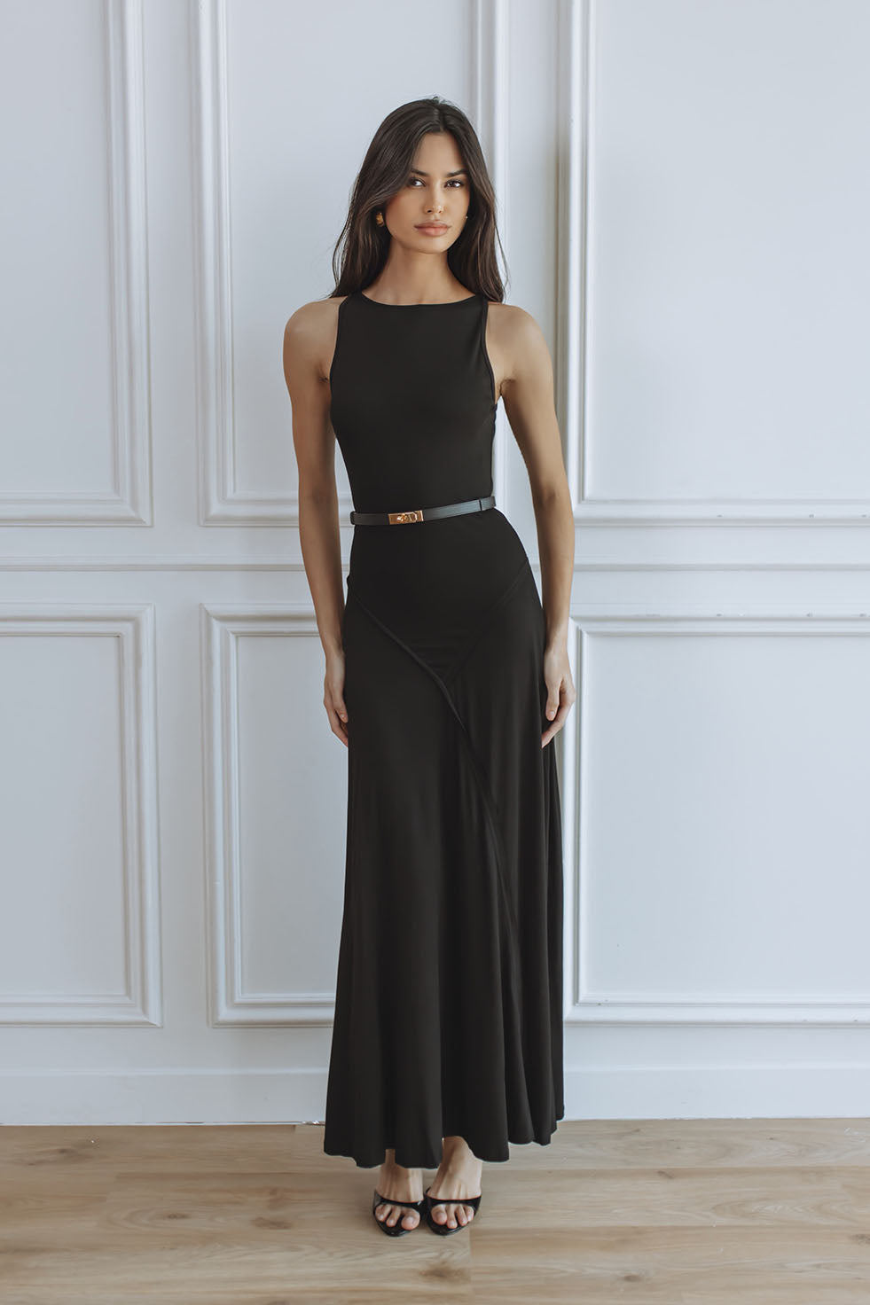 A woman modeling a sleeveless black maxi dress with a thin gold belt, standing against a white paneled wall on a light wood floor.