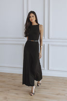 A woman modeling a black sleeveless maxi dress with a thin metallic belt at the waist, standing on a light wood floor against a white paneled wall.