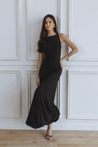 A woman modeling a sleeveless black maxi dress with a high neckline, standing against a white paneled wall on a light wood floor.