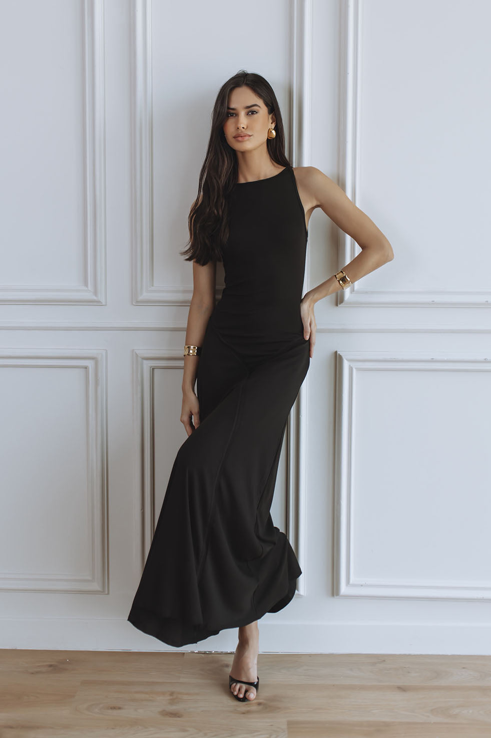 A woman modeling a sleeveless black maxi dress with a high neckline, standing against a white paneled wall on a light wood floor.