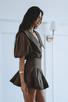 A woman wearing a dark brown short puff-sleeve dress with a wide collar and a flared skirt, standing in a bright white room with a wall lamp visible in the background.