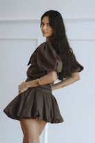 A woman modeling a dark brown puff-sleeve mini dress with a pleated skirt, posed against a clean white paneled wall.