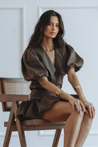 A woman sits on a wooden chair wearing a dark brown satin puff-sleeve mini dress with a deep V-neckline. She is accessorized with gold bracelets against a clean, light gray wall with white molding.