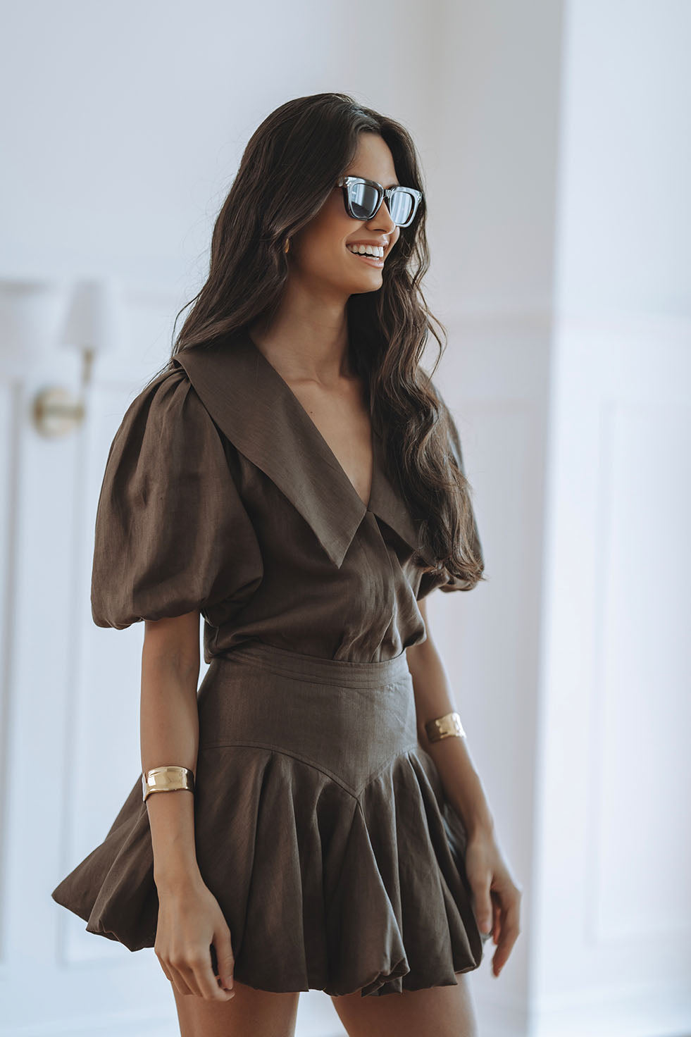 A woman wearing a brown short puff-sleeve dress with a large collar and pleated skirt, styled with sunglasses and gold bracelets, standing in a bright white room.