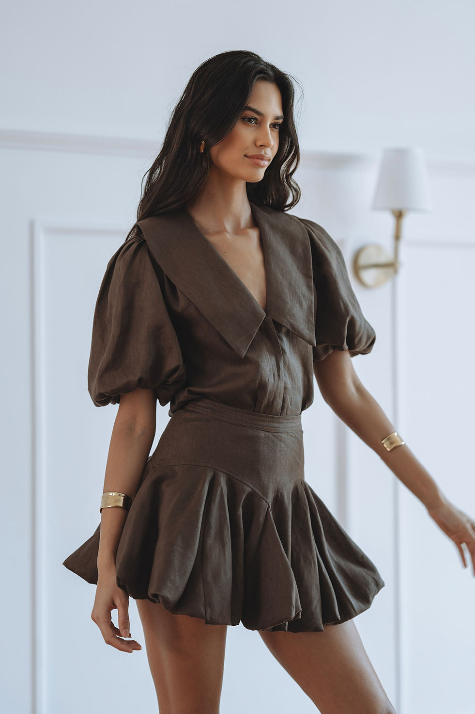 A woman modeling a brown short puff-sleeve dress with a wrap-style V-neck and pleated skirt, posed in a bright indoor setting with a wall lamp in the background.